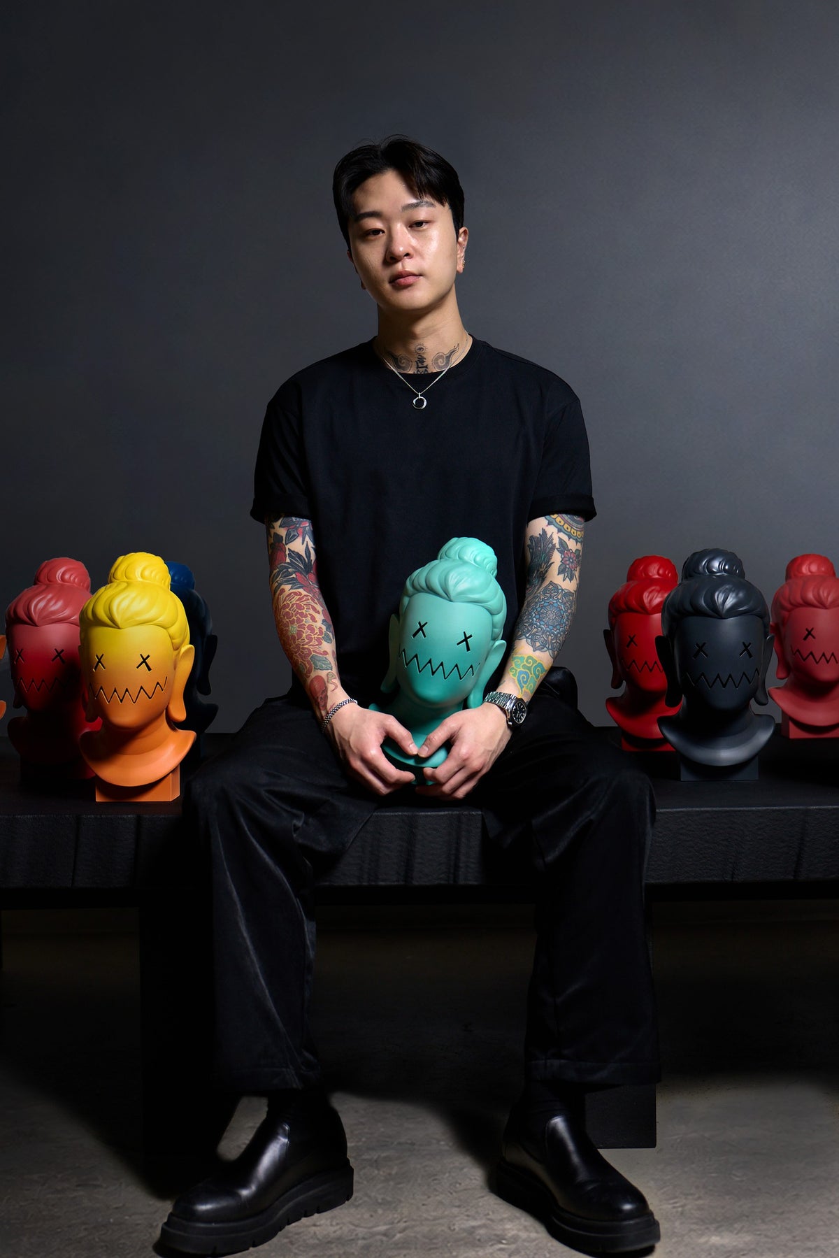 PittaKKM seated on a bench holding a teal sculpted head, with rows of his coloured heads either side