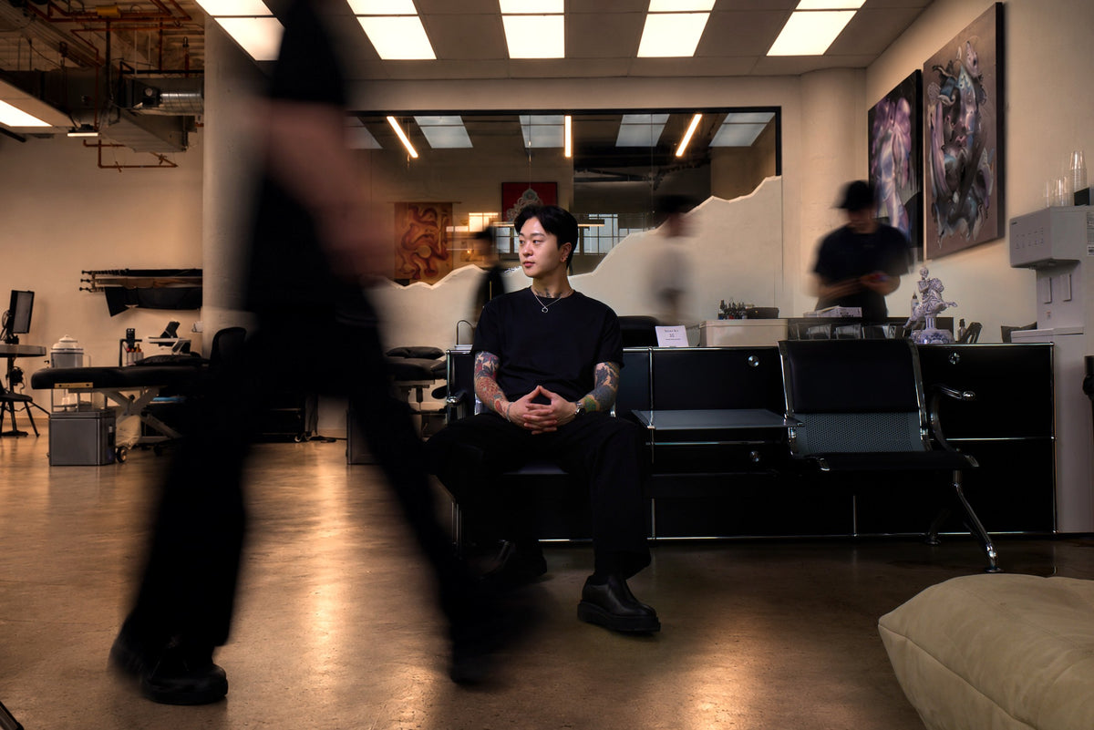 PittaKKM sitting still on a bench in his studio while people move past him in a blur