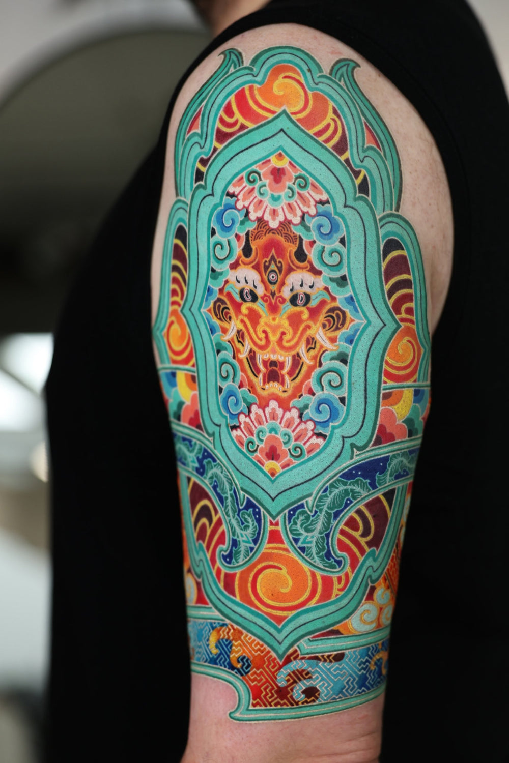 PittaKKM upper arm tattoo, a fanged three-eyed guardian face in a teal frame surrounded by lotus, cloud and brocade pattern