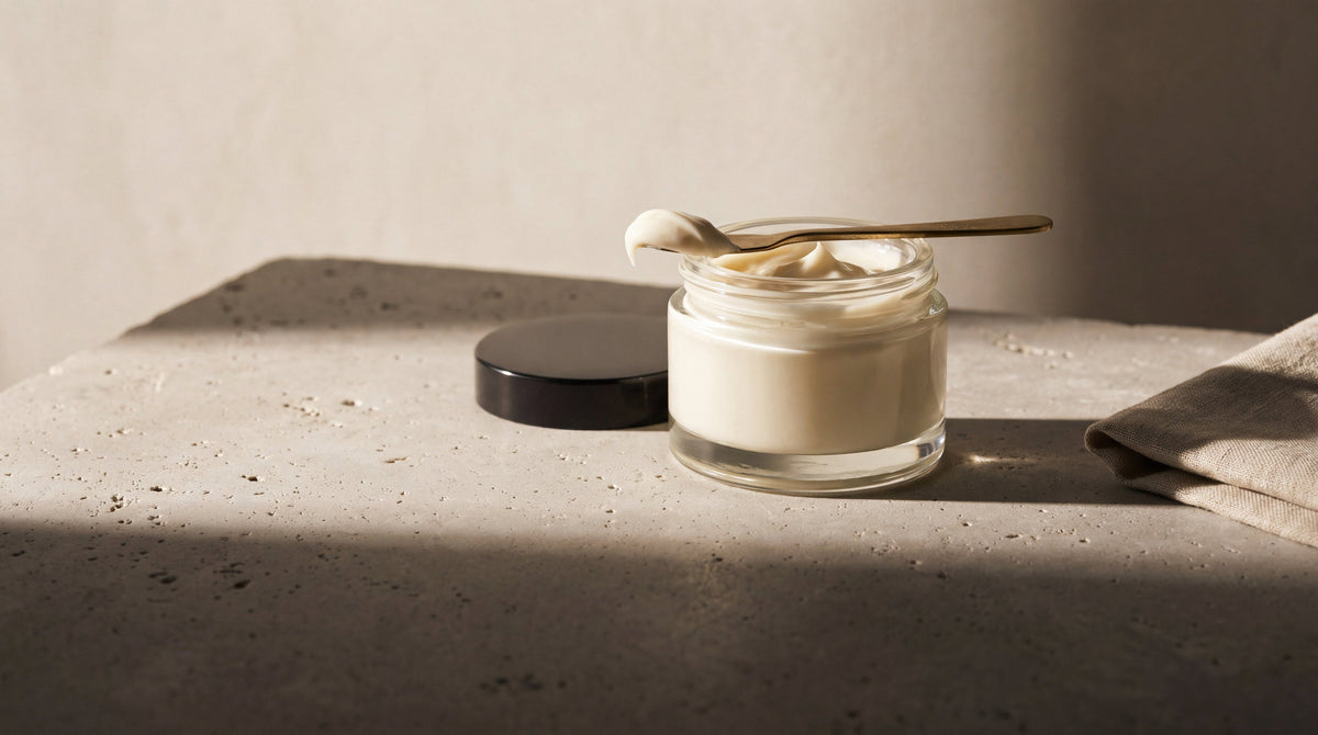 A glass jar of rich ivory cream with a brass spatula on travertine, choosing the best cream for a new tattoo