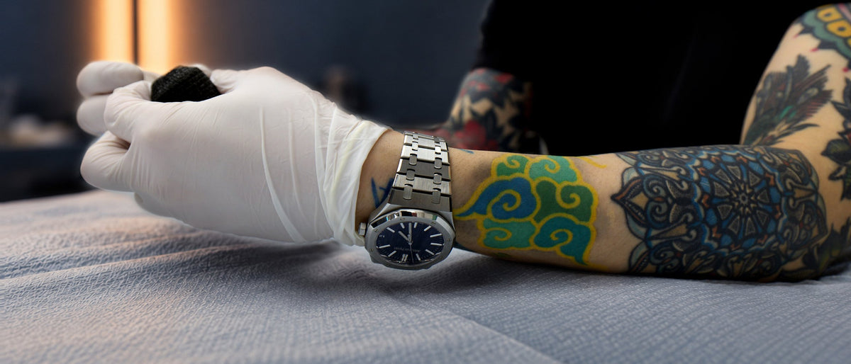 PittaKKM's gloved hands holding a tattoo machine at the bed, his own tattooed forearm beside them