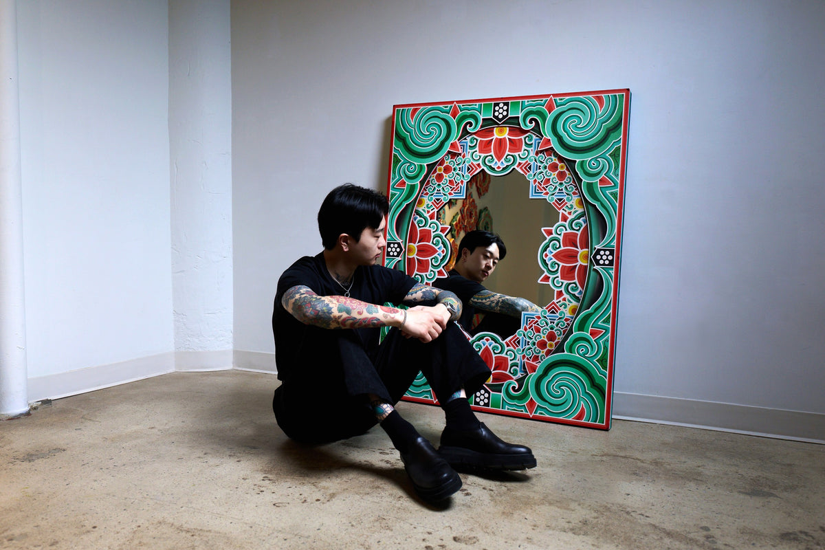 PittaKKM sitting on the floor beside a large dancheong frame with a mirror at its centre, turned towards his reflection