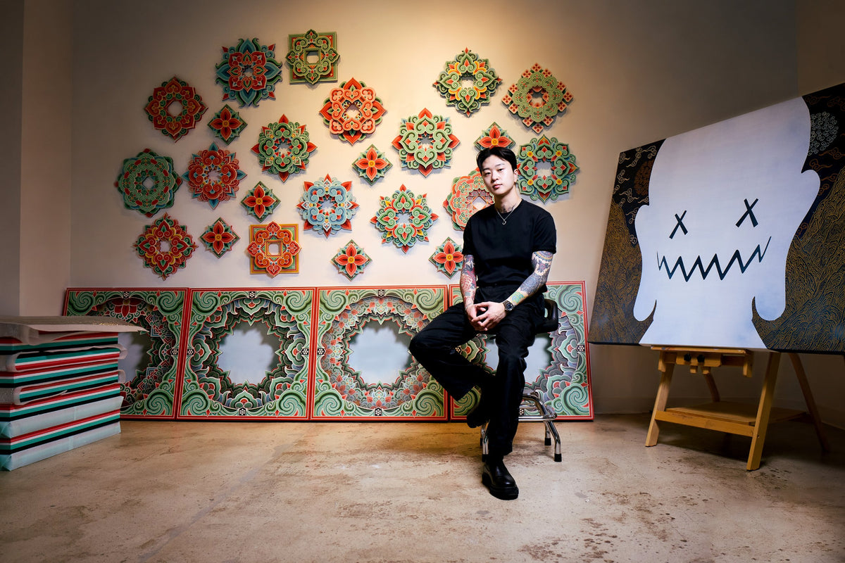 PittaKKM seated in his studio in front of a wall of dancheong panels, a large painted canvas on an easel beside him
